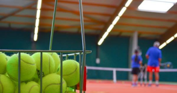 Bucket of tennis balls in an indoor tennis court. People play tennis in the background