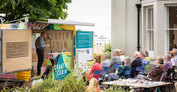 A person stands delivering a talk in the creative cabin to a seated audience in the Thelma Hulbert Gallery garden