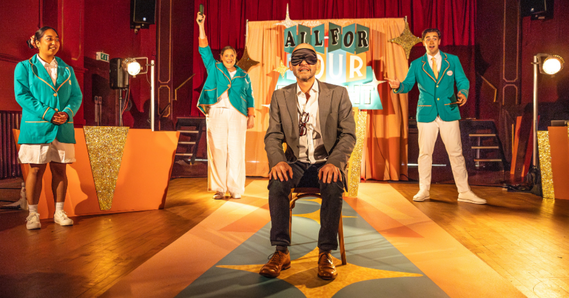 All For Your Delight. A person sits on a chair on stage, blindfolded. Three people are stood behind