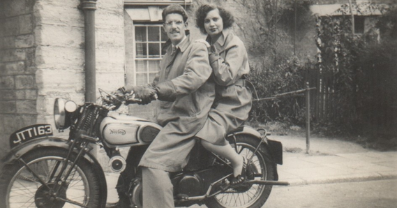 Black and white. Two people on a motorbike with a building in the background