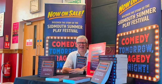 Paul Smith sat at a table with a pile of books and banners 'now on sale! Discover the story of the Sidmouth Summer Play Festival'