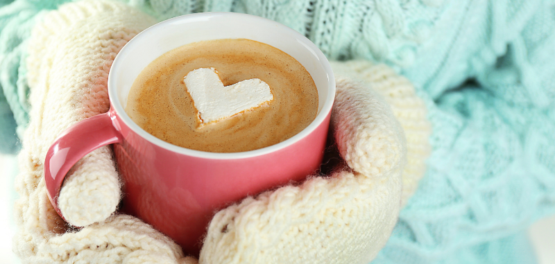 Person wearing gloves holding mug of coffee with heart shape decoration on top of coffee