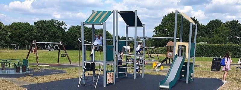 Cobham Downside play area. Children playing in a park with a climbing frame, swings, a slide, and a roundabout.