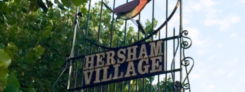 Metal gate with a robin design and the word Hersham set amongst green bushes 