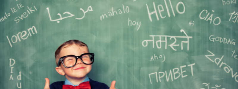 Smiling child with oversized glasses in front of a chalkboard covered with the words hello in different languages