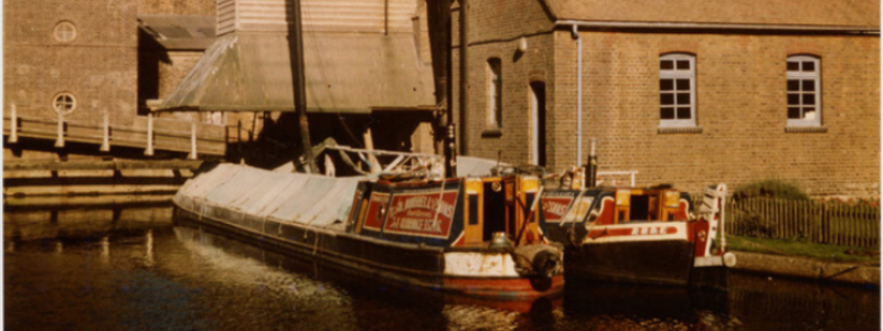 Sepia image of two canal boats on the river outside brick buildings