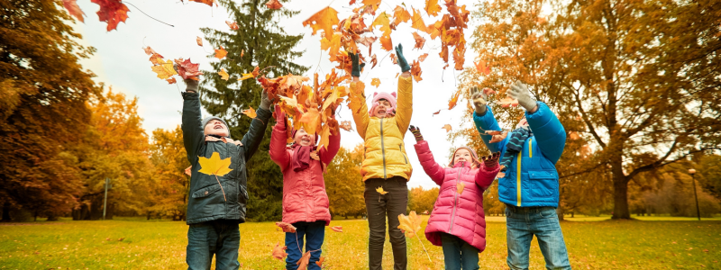 Five children wearing coats throwing autumn leaves in the air in a park setting