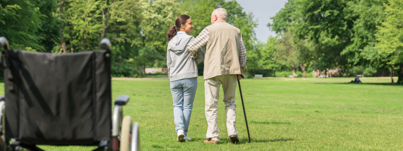 Wheelchair in foreground and elderly gentleman accompanied by carer, walking away