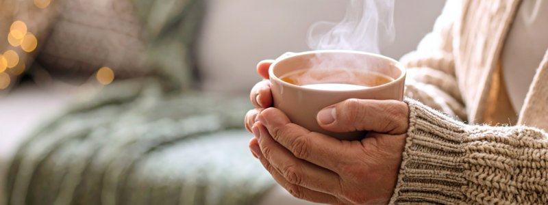 A lady, wrapped in a cream cardigan and holding a cup of warm tea.