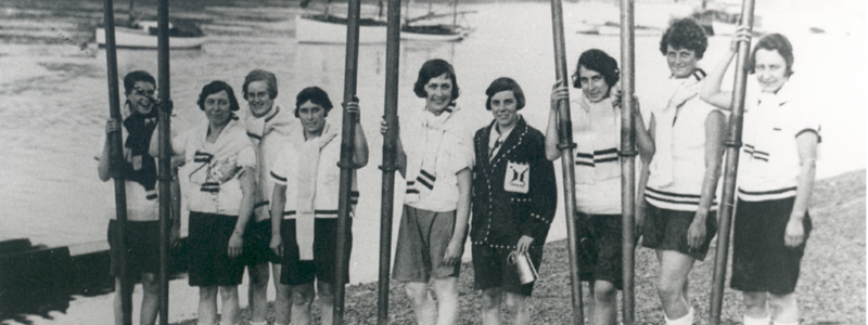 Black and white photo of nine women holding oars dressed in rowing outfits standing in front of a river with boats
