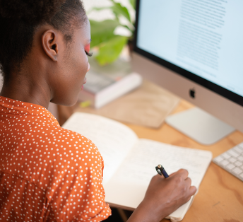 person writing in a note book while sat in front of a computer screen