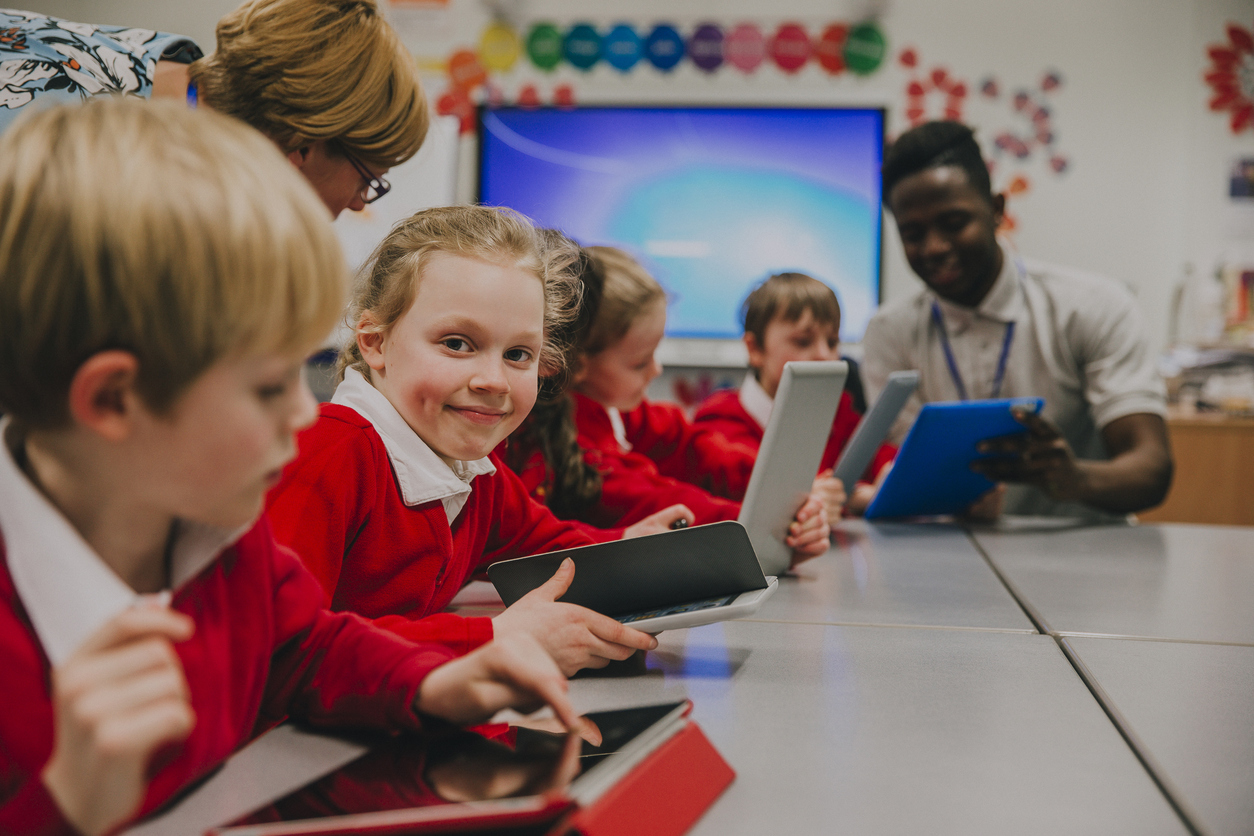 Children  sat at a long desk in a classroom in red uniforms working on ipads with their teacher while a little girl looks at the camera. 
