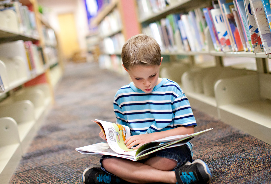 A boy sits cross-legged on a library floor, absorbed in the book sitting on his lap