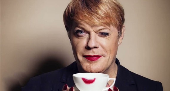 A close up of Eddie Izzard, holding a cup of tea with lipstick marks on the rim and winking at the camera