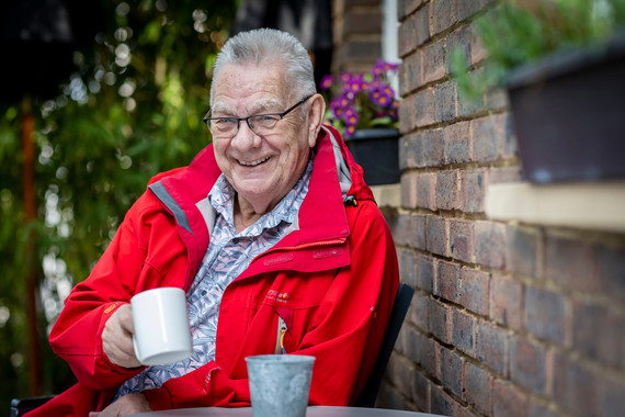 A older man in a bright red jacket drinks a cup of tea and smiles at the camera