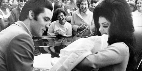 A black and white photo of Elvis Presley and his wife Priscilla holding their baby with an adoring crowd in the background