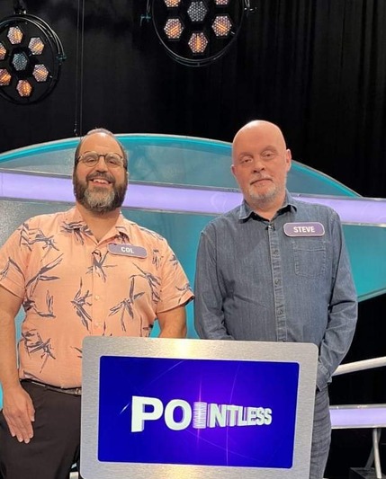 Two men stood in in front of a lectern, with the word 'Pointless' written on it, in a TV studio.