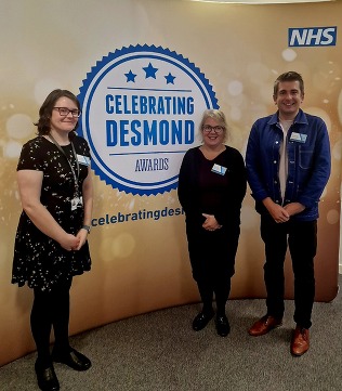 Three people stood in front of a sign which says 'Celebrating Desmond awards'.