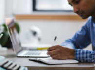 Man writes notes in notebook while working on laptop.