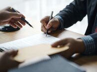 The hands of two people sitting on opposite sides of a table, holding a pen each and examining a document on the table.