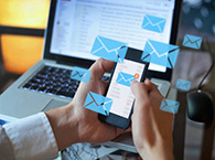 An office worker sends an email from their phone, with envelope icons floating around the phone and a laptop open in the background.