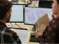 On the left, a male caseworker in a plaid shirt looks at a mapping screen on his computer, while a female caseworker to his right offers advice.