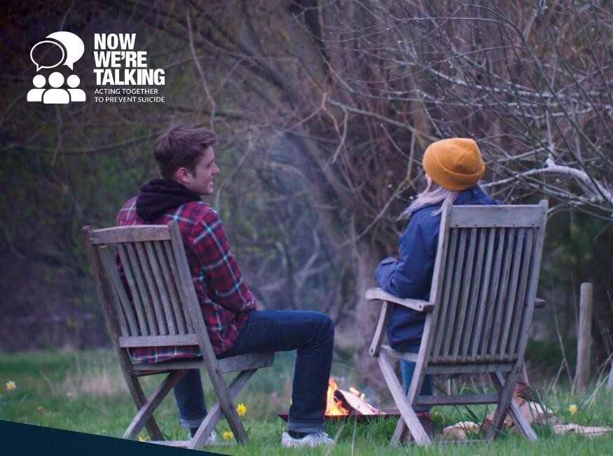 Two people sat on wooden chairs outside, talking to each other