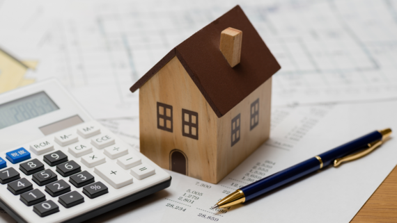 a small wooden house next to some bills, a calculator and a pen