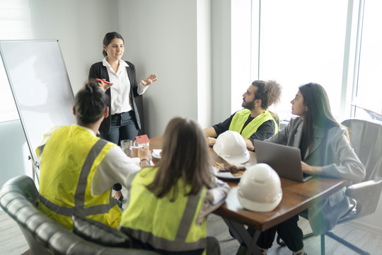 Group of managers sat around a table listening to a women standing in front of a flip chart