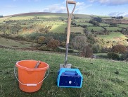 Orange bucket, spade, and blue farming equipment on grassy hill with countryside in the background