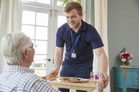 A smiling younger man is handing a tray of dinner to an older man in a sunny care home.