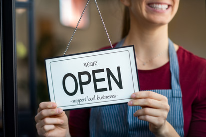 A person behind a shop door holding a sign that says 'We are open. Support local businesses.'
