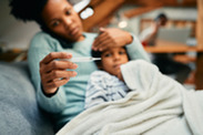 A black woman cuddled up on the sofa with their sick child holding a thermometer and their child's forehead