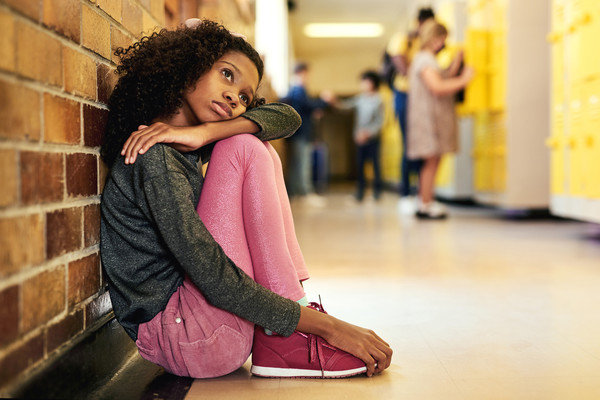 A young Black girl sat in a school corridor looking sad and resting her head on her knee