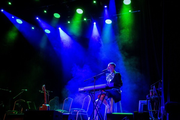 A queer artist Mark T Cox playing the piano on a stag in blue lighting