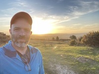 A man wearing a blue cap, standing in front of a sunset