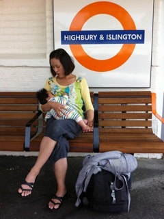 A woman sitting on a bench breastfeeding her baby, in front of a sign that says Highbury and Islington