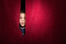 A young boy poking his head out between two red curtains