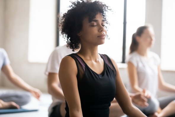 Photo of a lady sat in a meditation pose on the ground with eyes closed breathing deeply surrounded by others