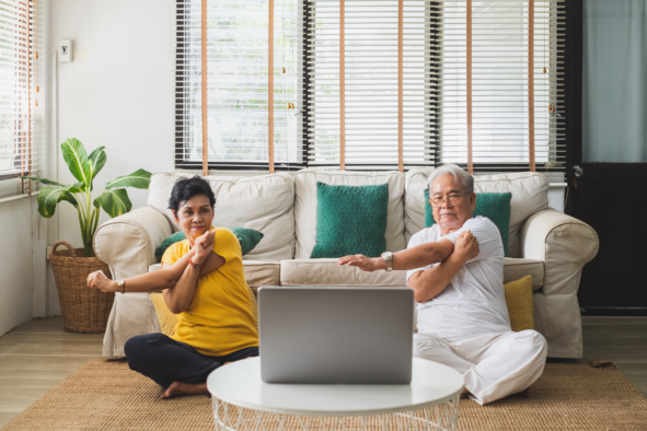 Two older people sat on their living room floor doing stretches looking at their laptop. They are doing an online exercise class