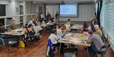 Groups of people sitting around tables in a hall watching three other people deliver a slideshow presentation at the front