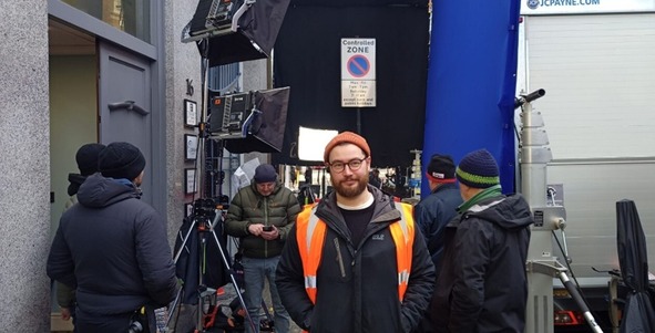 man in orange high vis vest standing in front of people and camera equipment