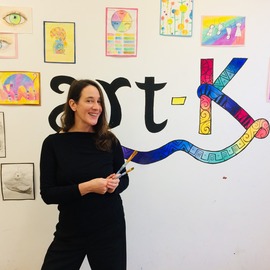 woman stands with paintbrushes in front of art-k wall