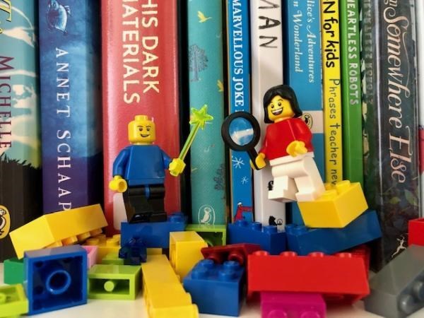 Two lego figurines with magnifying glasses in front of a shelf of books