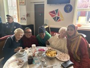 A group of older ladies from Be Caring sat at a table with food smile at the camera