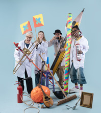 Four people in white coats in a line smiling at the camera and holding a random assortment of objects 