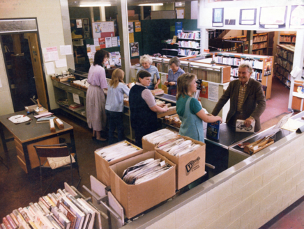 Image shows the inside of a library with lots of shelves of books, four staff behind the counter and three customers in front