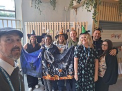 People stood in a row smiling a the camera. They are holding a halloween themed banner and wearing witches hats. 