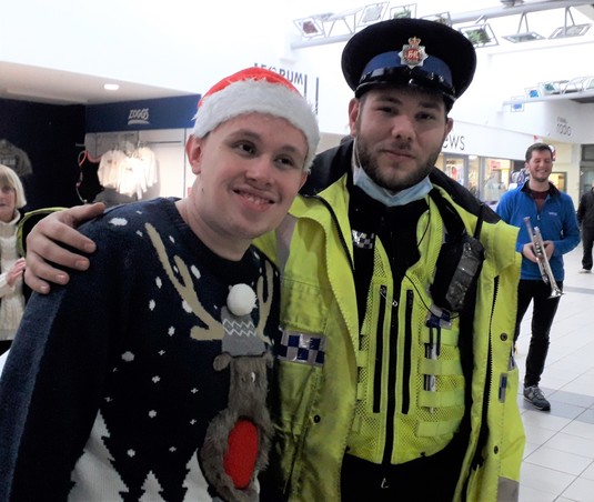 2 pale-skinned smiling men. 1 is a police officer in uniform, 1 is wearing a Christmas outfit
