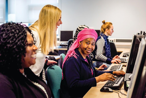 A diverse range of adult learners working on computers. Learners are smiling and their female tutor is supporting them